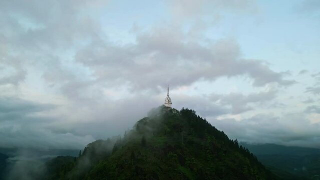 Drone circles Ambuluwawa Tower in Sri Lanka, rising above clouds on green mountain. Misty landscape around multi-faith monument. Aerial footage for travel, spirituality, nature, peace themes.