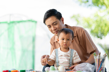 Asian father and his young son engaged in painting Easter eggs with watercolors, sitting on table...