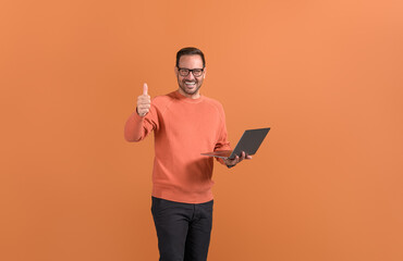 Portrait of smiling businessman with laptop showing thumbs up sign on isolated orange background