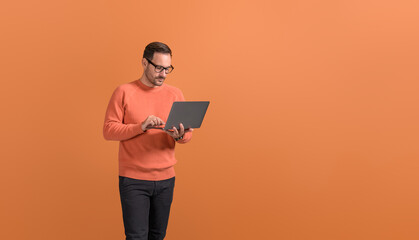 Portrait of focused male entrepreneur in glasses working over wireless computer on orange background