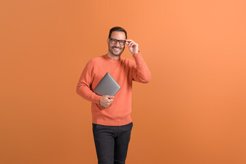 Portrait of confident businessman with laptop adjusting glasses while standing on orange background