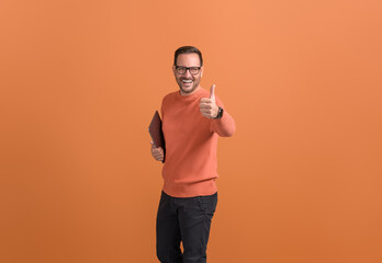 Portrait of cheerful handsome freelancer with laptop showing thumbs up sign over orange background