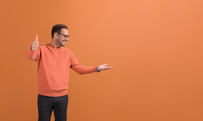 Satisfied happy male customer showing thumbs up sign and looking at empty palm on orange background