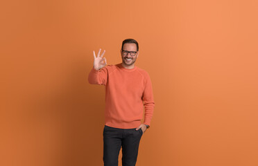 Portrait of satisfied male customer with hand in pocket showing OK gesture on orange background