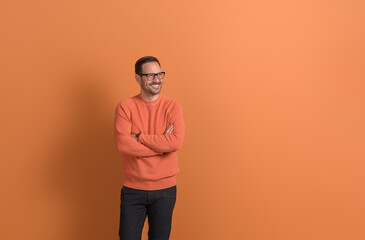 Smiling young businessman with arms crossed looking away and thinking on isolated orange background