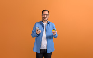 Portrait of cheerful male customer making mobile payment with credit card on orange background