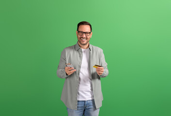 Smiling young entrepreneur paying bills online with credit card and mobile phone on green background