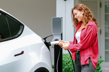 Modern eco-friendly woman recharging electric vehicle from home EV charging station. EV car...
