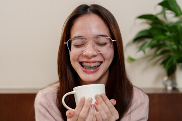 Youth holds white ceramic cup, closed eyes, wide smile revealing teeth with blue braces, hands...