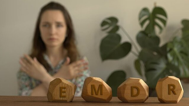 Letters EMDR written on irregular wooden blocks. A female is touching and tapping her shoulders in a blurred background. Eye Movement Desensitization and Reprocessing Psychotherapy Treatment concept.