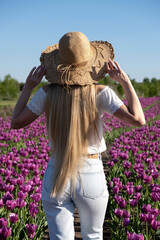 Young woman with long blond hair in blue jeans, white blouse and straw hat from behind in tulips field. Romantic scene of a beautiful woman in flower field on sunset. Tranquility and harmony.