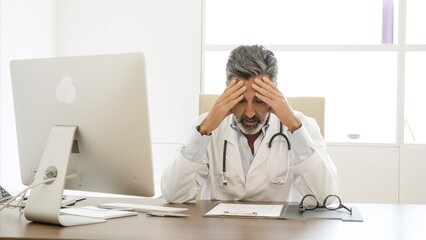 Stressed male doctor suffers from headache sitting at desk in clinic
