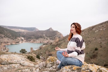 A woman is sitting on a rock overlooking a body of water. The scene is peaceful and serene, with the woman looking out over the water and taking in the view.