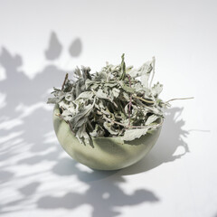 Dried Herbal Leaves in Ceramic Bowl on White Background. Bowl of dried herbs casting a soft shadow on a white background, evoking a sense of natural simplicity and calm. Homeopathic treatment.