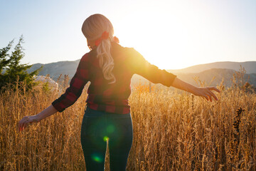 A blonde woman in a plaid shirt stands in a field of tall grass. happy woman outdoors at sunset on...
