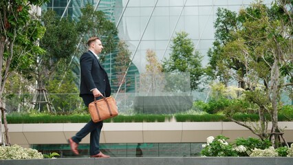 Side view of caucasian businessman hold suitcase and walking at modern building in green eco city. Professional male leader going to workplace at morning while wearing formal suit. Lifestyle. Urbane.
