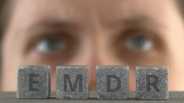 Eye Movement Desensitization and Reprocessing psychotherapy treatment concept. Letters EMDR written on grey stone cubes. Blurred woman's face engaging in side-to-side eye movements in the background.