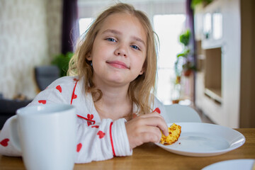 A cute little blonde girl in beautiful pajamas is sitting at a cozy table in the kitchen and having...