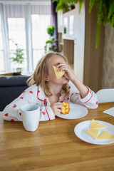 A cute blonde girl in beautiful pajamas is sitting at a cozy table in the kitchen and eating a delicious and healthy breakfast. The concept of proper and balanced nutrition.