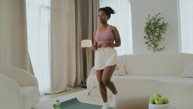 Young black woman with not a perfect figure in sportswear doing a workout at home in living room. She is a beginner in sports and uses laptop for online training.