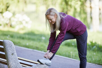 A sportive woman in her forties stretches her legs on a park bench during an early morning workout