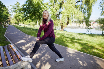 Mature woman performs stretching exercises on a park bench, enjoying a sunny day outdoors