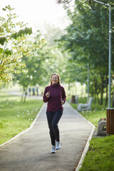 Cheerful middle-aged woman jogging in a sunlit park with lush greenery