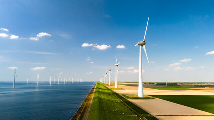 A row of towering wind turbines gracefully spin with the breeze next to a tranquil body of water in...