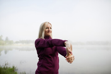 Mature woman stretching arms before lakeside exercise on a foggy day