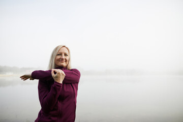Woman in her forties stretches outdoors on a foggy morning, dressed in a purple sports jacket