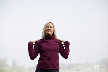 Photo Description:.Confident mature woman in a sporty outfit stretches her arms, enveloped in morning fog, poised and serene