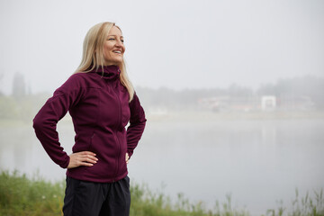 Smiling middle-aged woman confidently stands with hands on hips in fog, dressed in sportswear,...