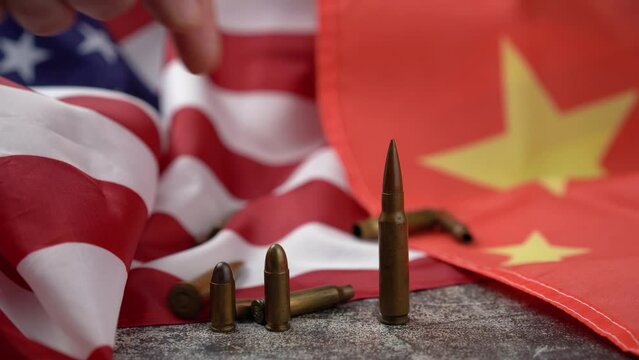 Unrecognizable person's hand pushing a large-caliber bullet placed in line with others, amidst an American and Chinese flag