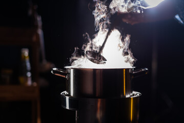 A boiling pot steams up as a chef uses a ladle to stir soup in a cooking pot in a restaurant.