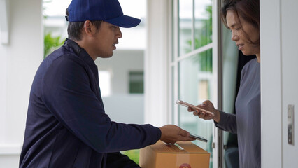 A delivery man in a cap presents a package to a woman at her doorstep, awaiting digital confirmation on her smartphone in a residential setting.
