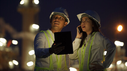Two construction engineers in hard hats and reflective vests using digital devices for night-time project coordination at an active construction site.
