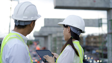 Two engineers in hard hats and reflective vests actively discuss and point at details in construction plans on a busy site.
