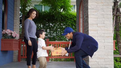 A friendly delivery man in a blue cap hands a package to a young child on the porch, with his mother watching, emphasizing a personal touch in service.
