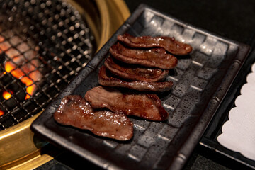 Grilled Ox Tongue on a Black Plate with Additional Cuts Cooking on a Gold Grill Over Charcoal Fire for an Authentic Japanese or Korean BBQ