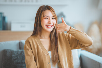 Satisfied asian woman smiling while looking at camera with thumbs up sign sitting on a couch in the...