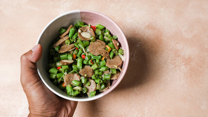 A hand holds a bowl containing stir-fried green beans with the addition of meatballs cut into pieces.