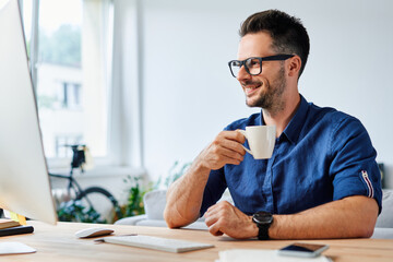 Smiling man drinking coffee looking at computer while working from home office