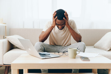 The Tired African American Freelancer Working on a Laptop in His Modern Home Office, Battling...