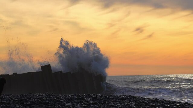 Wave hit large metal fence and rose high into air at sunset. Tsunami hits shore. Strong storm, wind and high big waves.