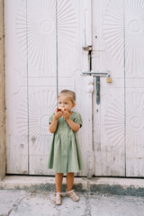 Little girl gnaws a big apple while standing near a wooden antique carved door