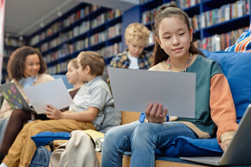 Portrait of young Asian girl using laptop in modern library lounge with group of children in backgroundwith copy space