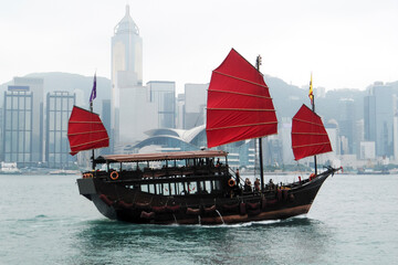 old Chinese boat (junk) under red sails; a group of tourists on a pleasure trip along the Victoria Strait (Hong Kong, China); skyscrapers; morning mist