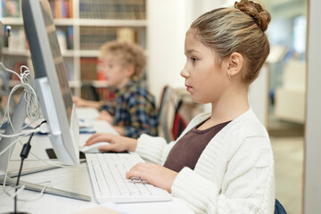 Side view portrait of serious young schoolgirl using computer in library during programming class for kidswith copy space - Powered by Adobe