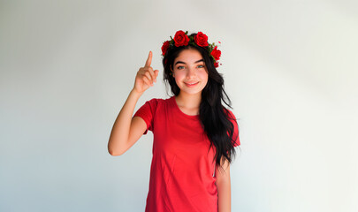 beautiful hispanic young woman with a crown of flowers on her head, wearing red t-shirt, pointing with her finger, isolated on white 