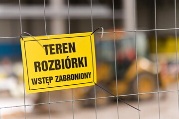 Znak Teren rozbiórki Wstęp zabroniony wiszący na płocie, w tle zarys koparki oraz budynku podczas budowy © Piotr